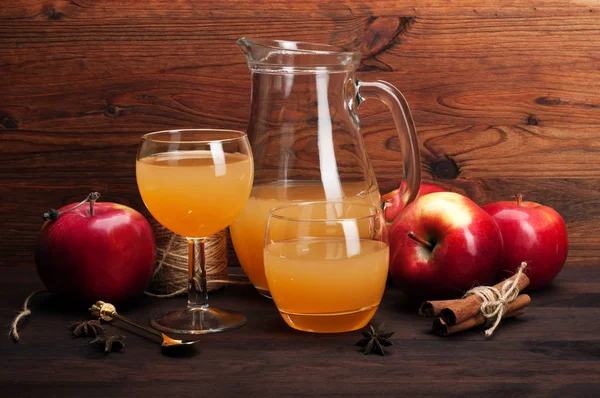 Apple cider, homemade in a glass jug and a glass, next to red apples and spices. Brown wood background.