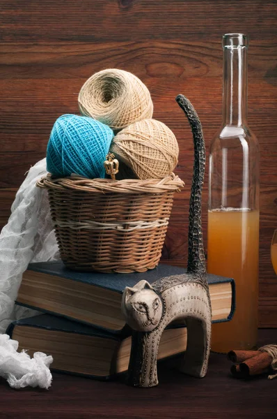 Still-life with cat statuette, a basket with beige and blue balls of thread, homemade cider on a brown wooden background.