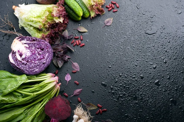 Vegetable background (wallpaper). Bright juicy purple and green vegetables and herbs on a black background with water drops. Red cabbage, purple onion, spinach, lettuce, cucumbers and basil. Space for text. Vegetarian, vegan concept