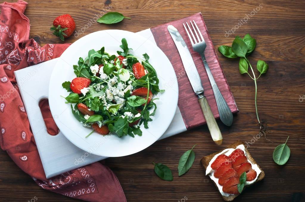 Salad with arugula, strawberries, blue cheese and olive oil on a white plate. Low-calorie diet healthy dish. Seasonal products. Close the sandwich with toast, low fat soft cheese and strawberries.