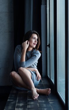 Gümüş elbisenin içinde giyinmiş kahverengi saçlı güzel genç kadın (kız) bir pencere üzerinde oturan ve pencereden dışarı bakarak. Gün ışığında çekim. Seçici odak üstünde yüz