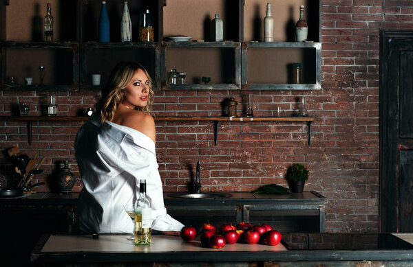 Beautiful young woman (girl) with white hair (blond), dressed in a white man's shirt, sitting at the kitchen table and looks over her shoulder into the frame. In the background kitchen loft-style