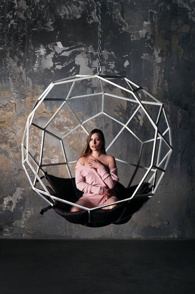 Beautiful young girl (woman) with a light auburn long hair in a pink cardigan sitting on the lap in a hanging chair on the background of a concrete wall. Hall in loft style.