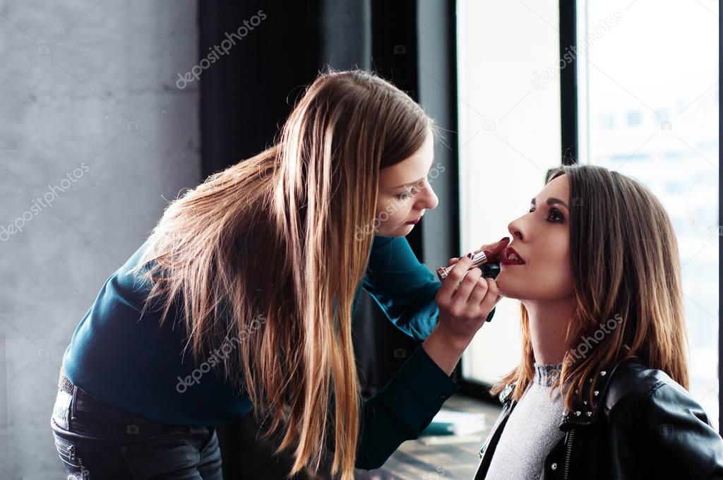 The process of make-up before taking a picture. The girl - makeup artist who paints a girl model with the help of dark red lipstick. The window in the background.