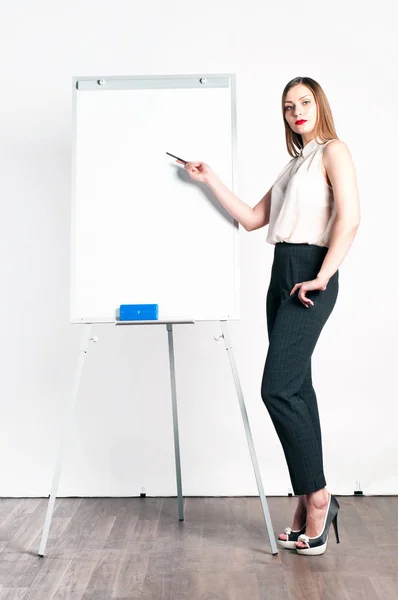 Beautiful young woman (girl) with long hair in a white blouse and short pants standing next to a flip chart and shows pointer on the flip chart. White background. Portrait in a profile in full growth.