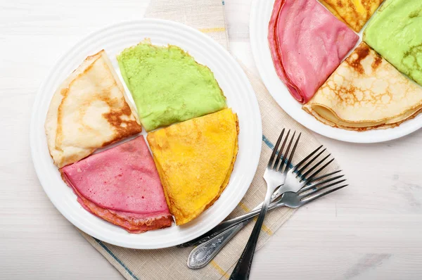 Panqueques multicolores hechos de leche, harina, azúcar y sal, con jugo de remolacha añadido, espinacas, zanahoria y cúrcuma en un plato blanco para la superficie de madera blanca. Original y hermosa comida casera — Foto de Stock