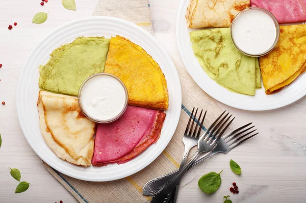 Crêpes multicolores à base de lait, farine, sucre et sel, avec du jus de betterave ajouté, des épinards, des carottes et du curcuma sur une plaque blanche pour une surface en bois blanc. Repas maison original et magnifique — Photo