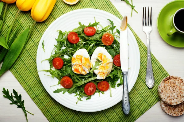 Arugula, 체리 토마토와 하얀 나무 바탕에 녹색 매트에 흰색 접시에 데친된 달걀에서 식이 저칼로리 샐러드. 근처 칼을 확인 하 고 있습니다. 건강 하 고 맛 있는 점심이 나 홈메이드 아침 식사. — 스톡 사진