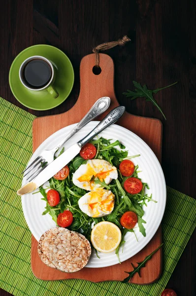 Dietary kalorisnål sallad från rucola, körsbärstomater och pocherade ägg på en vit platta på en grön matta på brun trä bakgrund. Det finns bestick i närheten. En frisk och läcker lunch eller hemlagad frukost. — Stockfoto