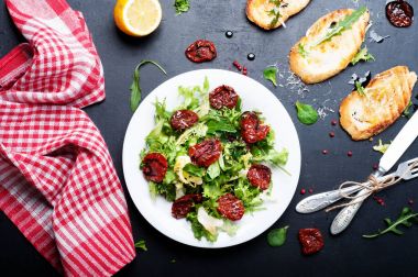Salata karışımı İtalyan otlar (buzdağı, esinti, mung fasulye, roka) ve güneş gelen karanlık bir yüzey üzerinde beyaz bir tabakta domates Kurutulmuş. İtalyan yemeği. Vejetaryen, vegan kavramı.