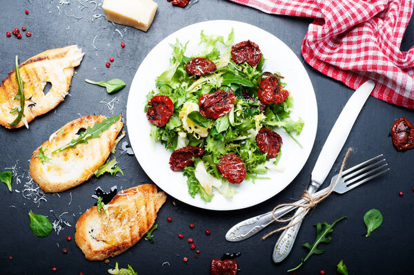 Salad from the mix of Italian herbs (iceberg, breeze, mung beans, arugula) and sun dried tomatoes on a white plate on a dark surface. Italian food. Vegetarian, vegan concept.