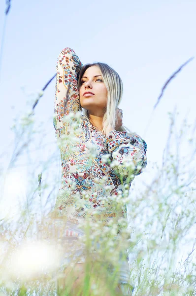 Ein schönes schlankes Mädchen (blond) in einer mehrfarbigen Bluse steht in Blumen vor dem blauen Himmel. Porträt bis zur Taille. Schießen im Freien bei Tageslicht — Stockfoto