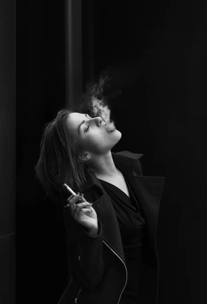 Una bella ragazza snella si alza di profilo, tiene una sigaretta in mano e lascia uscire il fumo. Foto in bianco e nero. Concetto retro. Ritratto alla vita — Foto Stock