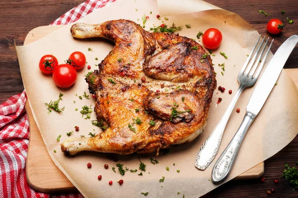 Stekt kyckling (kyckling) på bakplåtspapper, bredvid körsbärstomater, gaffel och kniv. Läckra kött stekt maträtt. — Stockfoto