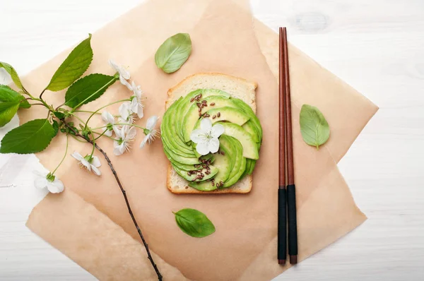 Sandwich with avocado and flax seeds on parchment. Next a branch with cherry blossoms and basil leaves. A useful low calorie ready dish. Vegetarian, vegan concept. Eastern cuisine — Stock Photo, Image