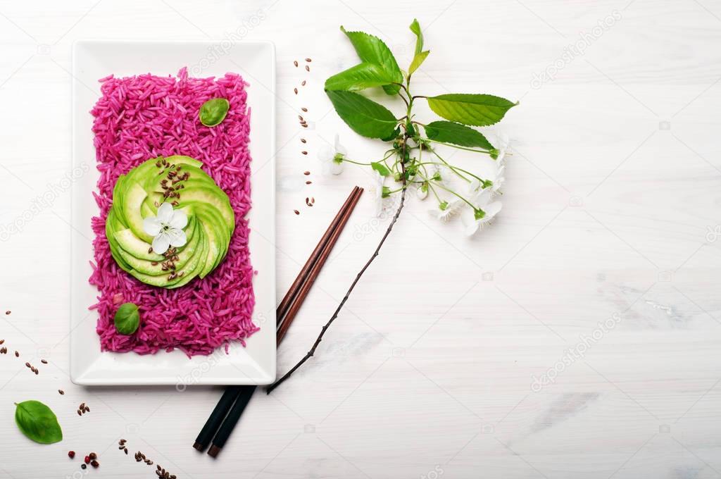 Pink rice with slices of avocado in plate on white wooden background. A branch with cherry flowers and Chinese sticks are next to it. A useful low calorie ready dish. Vegetarian, vegan concept. Place for text