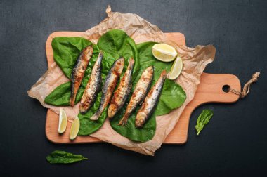 Karanlık bir yüzey üzerinde bir kesme tahtası üzerinde kızarmış sardalye (Deniz balığı). Ispanak yaprakları ve limon dilimlerini yakınında. Lezzetli ve sağlıklı vejetaryen ev yapımı Öğle Yemeği.