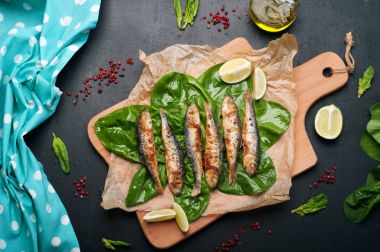 Karanlık bir yüzey üzerinde bir kesme tahtası üzerinde kızarmış sardalye (Deniz balığı). Ispanak yaprakları, zeytin yağı, çatal bıçak takımı ve kireç parçaları yanında. Lezzetli ve sağlıklı vejetaryen ev yapımı Öğle Yemeği.