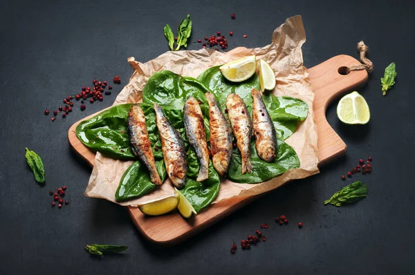 Жареные сардины (морская рыба) на режущей доске на темной поверхности. Рядом со шпинатом листья, специи и кусочки лайма. Вкусный и здоровый вегетарианский домашний обед . — стоковое фото
