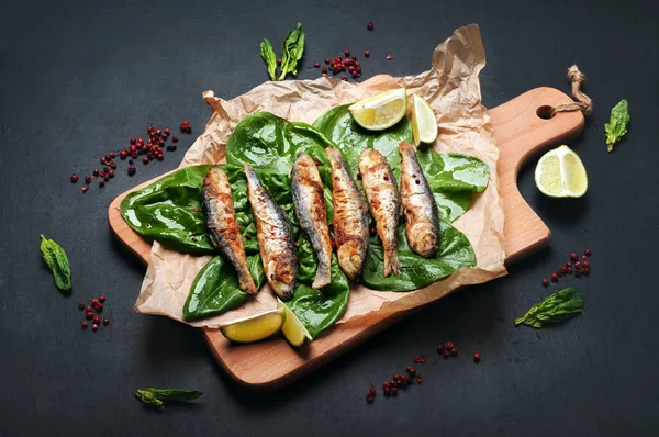 Smażone sardynki (ryby morskie) na pokładzie cięcia na ciemnej powierzchni. Obok liści szpinaku, przyprawy i kawałki limonki. Pyszne i zdrowe wegetariańskie lunch domowe. — Zdjęcie stockowe