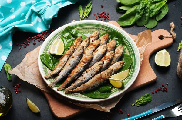 Sardinas fritas (pescado de mar) en un plato verde sobre una superficie oscura. Junto a las hojas de espinaca, aceite de oliva, cubiertos y trozos de lima. Delicioso y saludable almuerzo vegetariano casero . — Foto de Stock