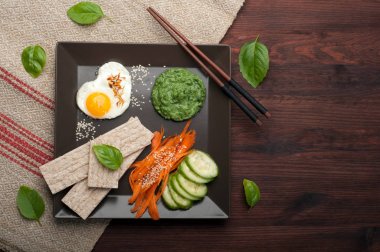 Onun sağlığı hakkında umurunda bir kişinin lezzetli ve sağlıklı bir kahvaltı kavramı. Sahanda yumurta, ıspanak püresi, diyet somun, salatalık ve havuç ahşap arka plan üzerine bir kare tabak içinde parçalar.