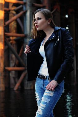 Güzel bir kız bir siyah deri ceket, beyaz bir T-shirt ve kot endüstriyel bir arka plan üzerinde duruyor. Gün ışığında, seçici netlik çekim. Yazarın renk düzeltme