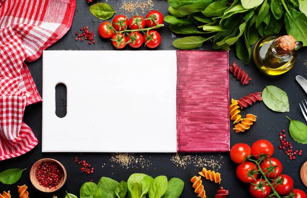 Background. Italian food. Multicolored pasta, tomatoes, spinach, basil leaves and olive oil on a dark surface. In the center of the image is wooden cutting board on which you can arrange dish or text — Stockfoto