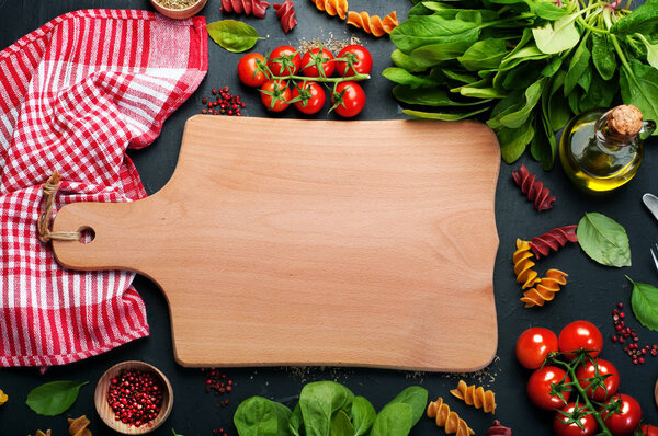 Background. Italian food. Multicolored pasta, tomatoes, spinach, basil leaves and olive oil on a dark surface. In the center of the image is wooden cutting board on which you can arrange dish or text