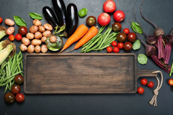 Vegetable background. A long brown wooden cutting board (tray), next to which lie fresh organic vegetables such as carrots, beets, asparagus beans, tomatoes, eggplant, on a dark surface. Place for text on the cutting board — Stock Photo, Image