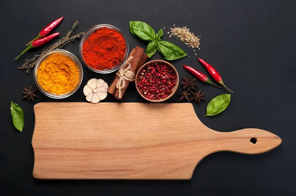 Bright spices, seasonings and herbs (turmeric, paprika, pink pepper, cinnamon, basil, thyme, anise and garlic) on a dark background. Near the light brown wooden cutting board (space for text) — Stock Photo, Image