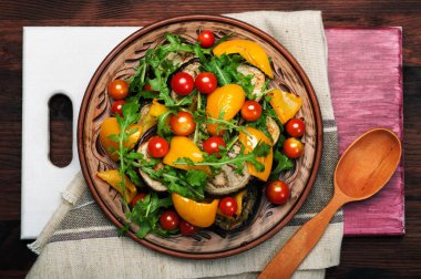 Mediterranean cuisine. Warm salad dish with baked pepper, eggplant, arugula leaves and cherry tomatoes on a brown clay plate. Vegan concept. Dietary low calorie food clipart