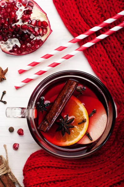 グリュー ワイン、リンゴ、オレンジ、スパイス、白い木製の背景の上に蜂蜜のスライスとガラスのコップ。近くに赤いスカーフ、ストロー、ザクロの半分。冬クリスマスと新年を飲む — ストック写真