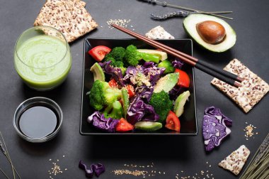 Brokoli, mor lahana, domates ve tatlı sosu ve karanlık bir arka plan üzerinde siyah bir kare tabak içinde zeytinyağı ile Avokado salatası. Yeşil smoothies ve soya sosu. Sağlıklı Diyet beslenme kavramı