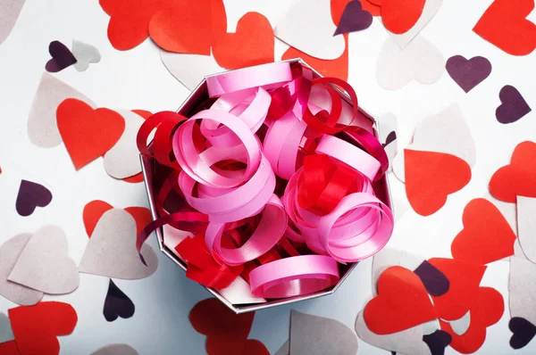 Festive decorations. Red and pink ribbons in a box on a white background. Nearby lay colored paper hearts. Celebration concept. — Stock Photo, Image