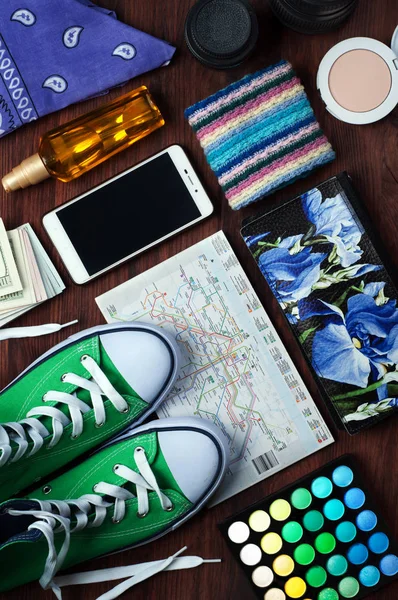 Let\'s travel! Objects for travel. Green sneakers, city map, cosmetics, purse, shawl, telephone, a fragment of bills and interchangeable lenses for the camera on a brown wooden background.