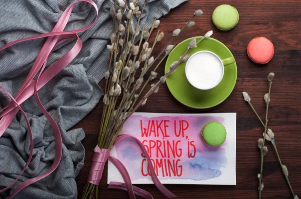 Coffee with whipped milk in a green cup on brown wooden background. Near the willow branches with a pink bird (decoration) and sweets (macaroons). 'Wake up, spring is coming' concept — Stock Photo, Image