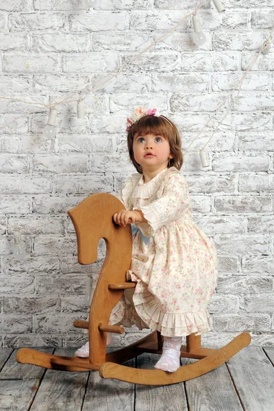 Cute child (a girl of two years old) in a flower dress and white pantyhose sits on a wooden horse against a white brick wall. Scandinavian style. Emotion `slight surprise`