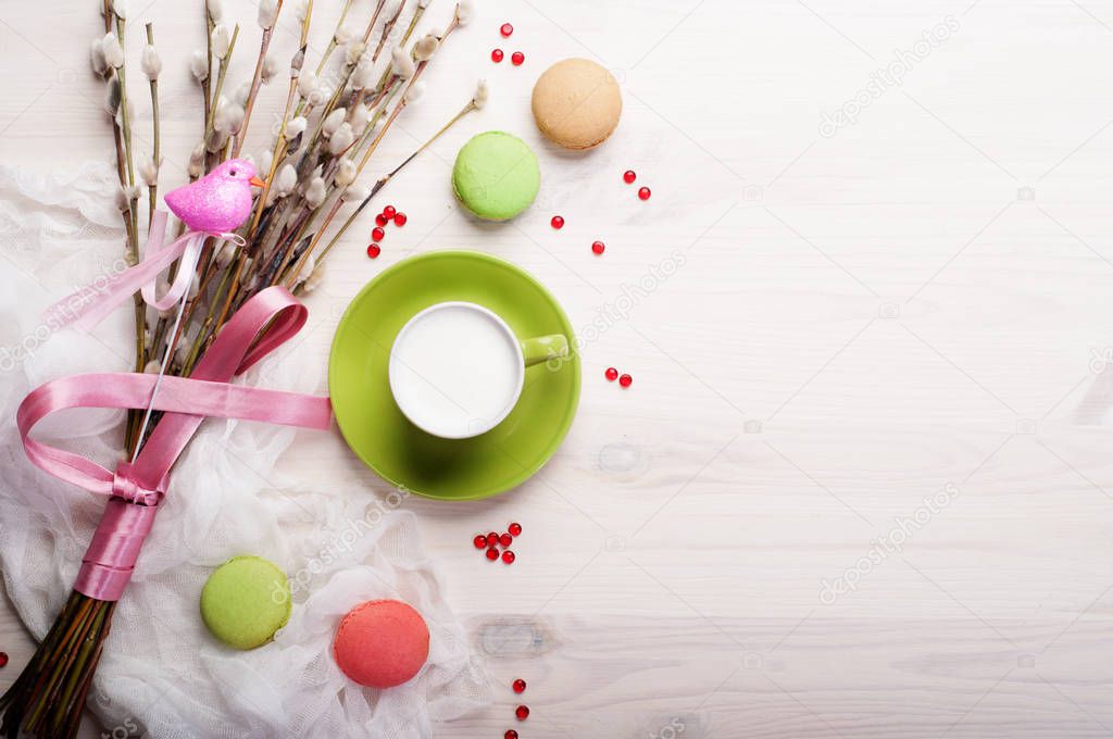 Coffee with whipped milk in a green cup on a white background. Near the willow branches with a pink bird (decoration) and sweets (macaroons). Place for text