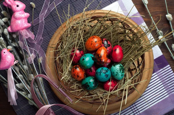 Pink, orange and green quail eggs on the hay in the basket. Near pussy-wilows and Easter decor (pink bird and bunny).  Brown wooden background.  Concept Easter food — Stock Photo, Image