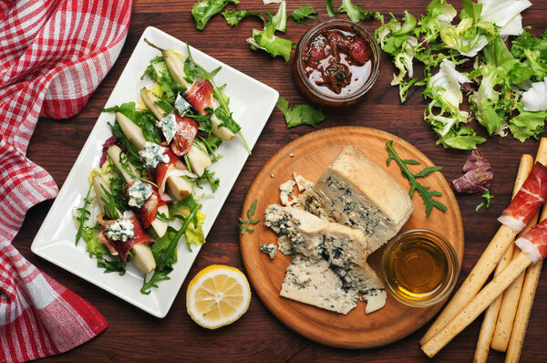 A large piece of blue cheese on a round cutting board. Dark wooden board. Next to olive oil, pear with dried meat and a mix of salad, lemon. Mediterranean style