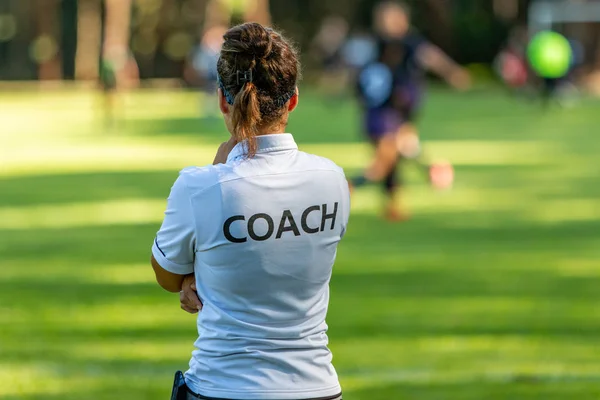 Bir bayan spor koçunun, takımının açık hava futbol sahasında yarışmasını izlemesi.