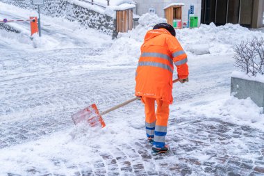 Açık turuncu ceketli yol temizleme işçisi. İsviçre, St. Moritz 'deki bir sokaktan kar temizliyor.