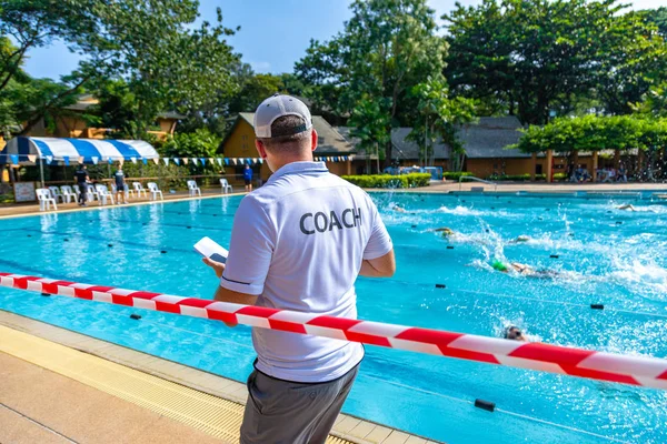 Back view of a male coach in white COACH shirt watching his swimmers racing at an outdoor swimming pool on a bright sunny day