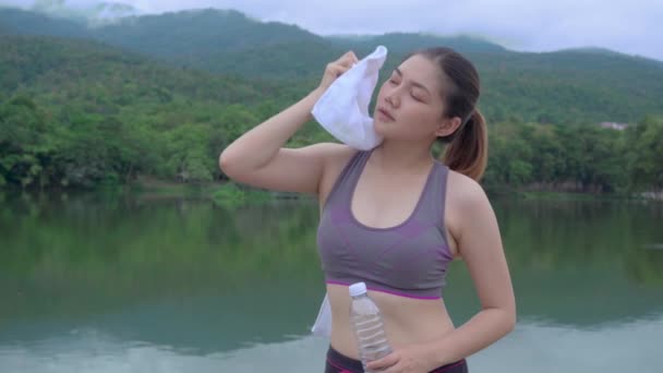 年轻健康的亚洲女人在一个有着湖水和高山美景的室外自然公园里 在早晨跑步时 停下来喝水 擦拭汗水 这是一种健康的生活方式 — 图库视频影像