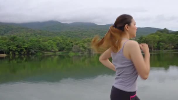 年轻健康的亚洲女人在当地的一个公园里进行晨练时跑步 带着湖景和山景 有着健康的生活方式理念 — 图库视频影像
