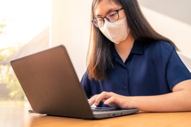 N95 maskeli genç bir Asyalı kadın COVID-19 virüsü yayılırken bilgisayarında çalışıyor. Sağlıklı bir konsept.