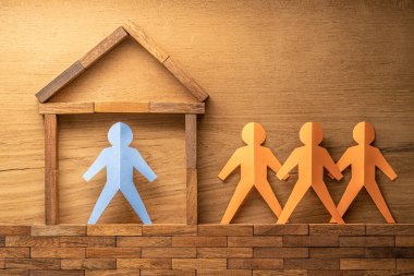 Tahta bir bloğun içindeki mavi kağıt kesiği insan figürü ve ahşap arka plandaki ahşap evin dışındaki üç turuncu kağıt kesme figürü COVID-19 virüs salgını sırasında evde kalıyor.