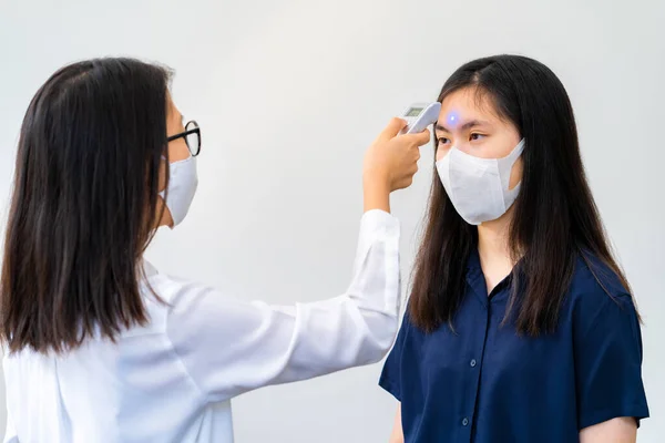 A young woman wearing face mask using handheld temperature checker to check temperature for another young woman wearing face mask, health concept