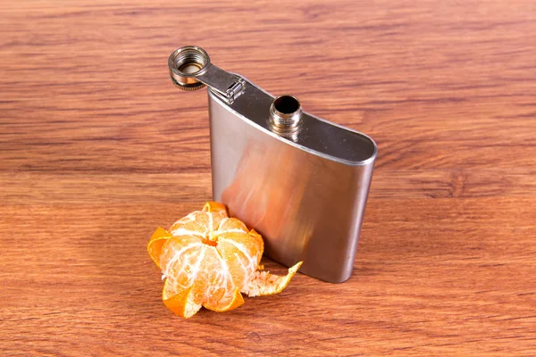 Стальная фляжка со спиртом на столе, вместе с ломтиком мандарина. Алкогольная зависимость . — стоковое фото